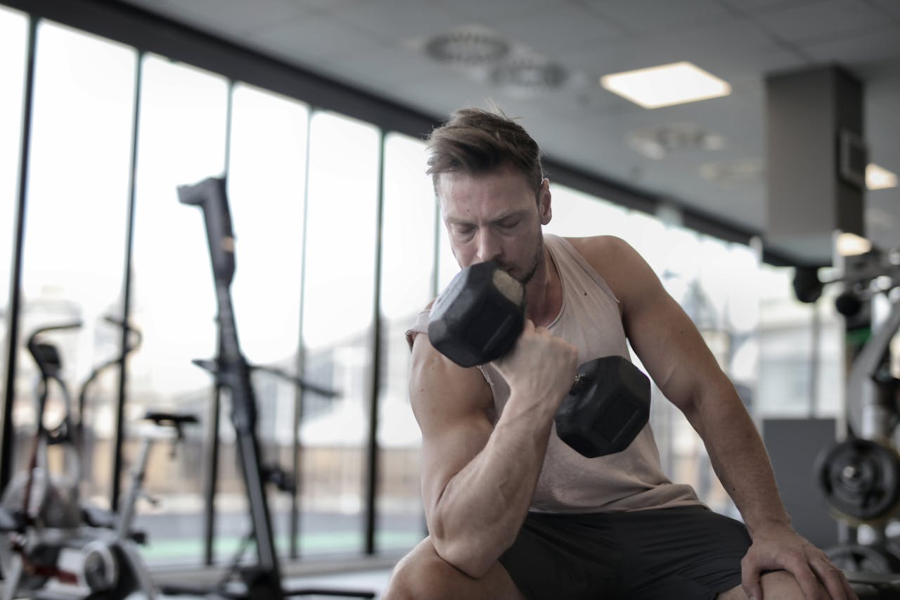 Offerings Low angle of powerful male athlete in sportswear performing exercise with heavy metal dumbbell while sitting on bench during workout in modern gym