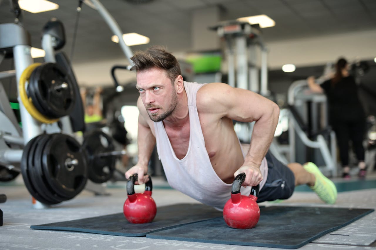 Home Low angle of strong sportsman making effort and pushing up on metal kettlebells during workout in modern gym and looking away